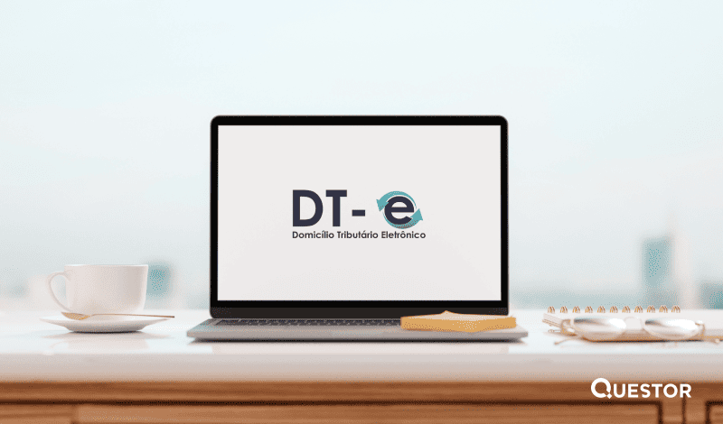 Imagem de um computador portátil exibindo o logo do Domicílio Tributário Eletrônico (DT-e) em uma mesa de escritório moderna, com uma xícara e objetos de papel ao redor, simbolizando serviço digital de gestão tributária.