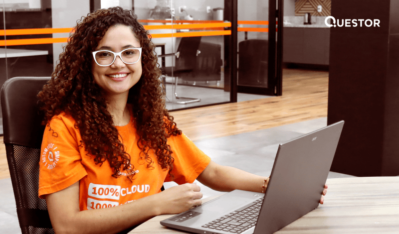 Mulher sorridente com óculos brancos, camiseta laranja com logo de nuvem e desktop no escritório moderno, representando trabalho remoto ou tecnologia.