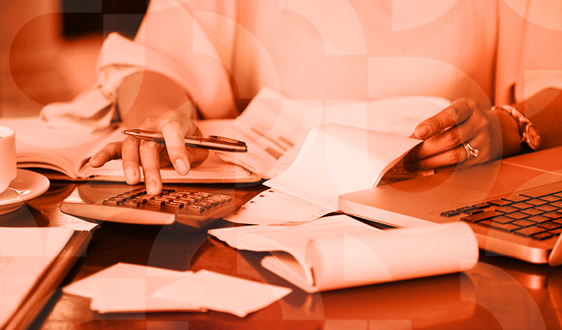 Pessoa trabalhando com papéis, laptop, caneta e calculadora em ambiente de escritório, representando trabalho e organização.